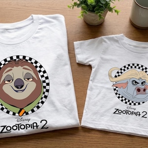 two white t-shirts with cartoon animal characters from the Disney movie "Zootopia 2" printed on them. The t-shirts are placed on a wooden surface, along with a coffee mug and a potted plant.
