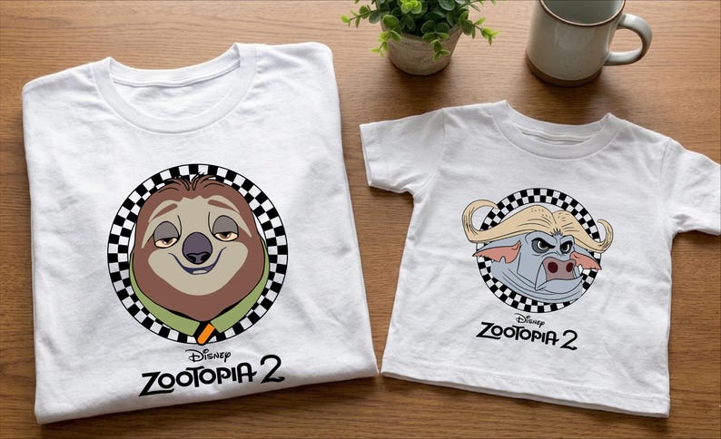 two white t-shirts with cartoon animal characters from the Disney movie "Zootopia 2" printed on them. The t-shirts are placed on a wooden surface, along with a coffee mug and a potted plant.