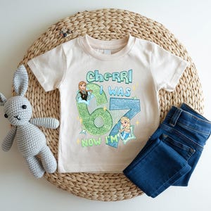 A white t-shirt with a graphic design, a gray crocheted stuffed rabbit, and a pair of blue jeans are arranged on a woven placemat.