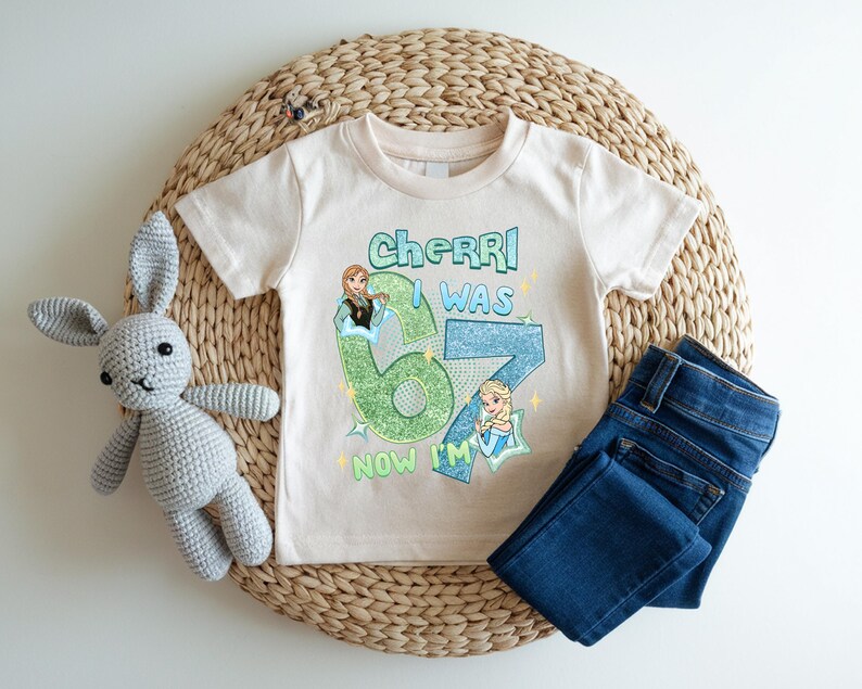 A white t-shirt with a graphic design, a gray crocheted stuffed rabbit, and a pair of blue jeans are arranged on a woven placemat.