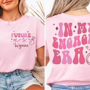 May include: Pink t-shirt with the text "Future Mrs. Wynne" and a ring on the front. The back of the shirt has the text "In My Engaged Era" in a retro font with a ring.