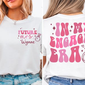 May include: Two white t-shirts with pink text and graphics. The front shirt says "Future Mrs. Wynne" with a pink ring and diamond graphic. The back shirt says "In My Engaged Era" with a pink ring and diamond graphic.