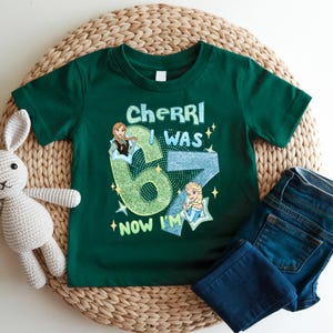A green t-shirt with a graphic design, a crocheted stuffed rabbit, and a pair of blue jeans are arranged on a woven mat.