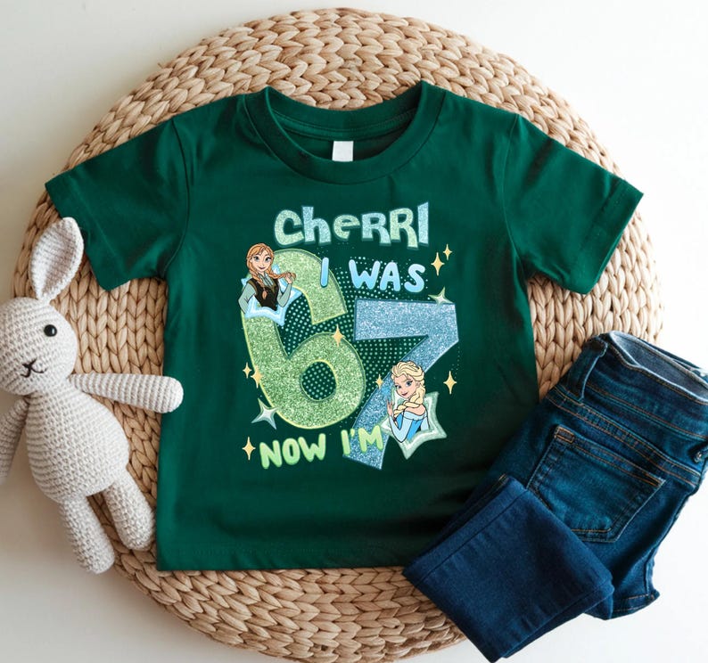A green t-shirt with a graphic design, a crocheted stuffed rabbit, and a pair of blue jeans are arranged on a woven mat.