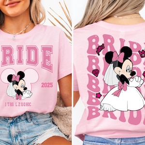 Può includere: Maglietta rosa con un design da sposa con un cartone animato di Topolino in abito da sposa. La parte anteriore della maglietta dice "BRIDE EST. 2025" e la parte posteriore della maglietta ha un design ripetitivo di "BRIDE" con Topolino con un abito da sposa.