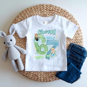 A white t-shirt with a graphic design, a crocheted stuffed rabbit, and a pair of blue jeans are arranged on a woven placemat.