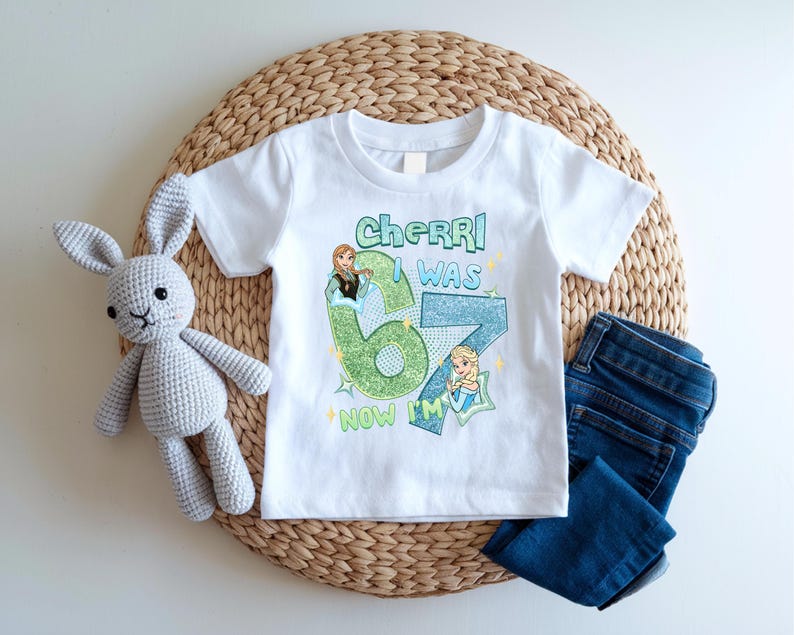 A white t-shirt with a graphic design, a crocheted stuffed rabbit, and a pair of blue jeans are arranged on a woven placemat.