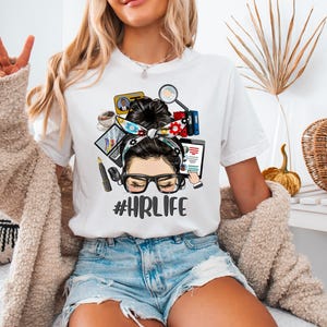 Op de afbeelding: Wit T-shirt met een afbeelding van een vrouw met een bril en een hoofdband, omringd door HR-gerelateerde iconen. De tekst "#HRLIFE" staat onder de afbeelding. Het haar van de vrouw is in een knot gestyled. Het shirt wordt gedragen met een spijkerbroek.