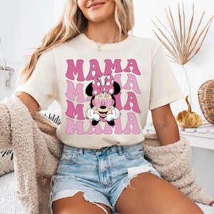 May include: A white t-shirt with a pink and white graphic of Minnie Mouse wearing heart-shaped sunglasses and the text "MAMA" repeated in a stacked pattern.