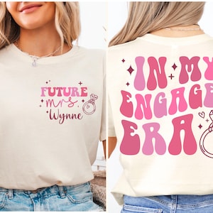 May include: A white t-shirt with pink and red text that says "Future Mrs. Wynne" on the front and "In My Engaged Era" on the back. The text is in a retro font style with a ring and diamond graphic.