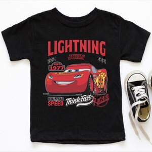 A black t-shirt with a graphic design featuring a red car and the text "Lightning John" along with other text and logos related to the Disney Pixar movie Cars.