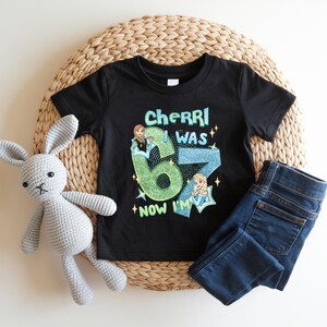 A black t-shirt with a graphic design, a gray crocheted rabbit, and a pair of blue jeans are arranged on a woven mat.