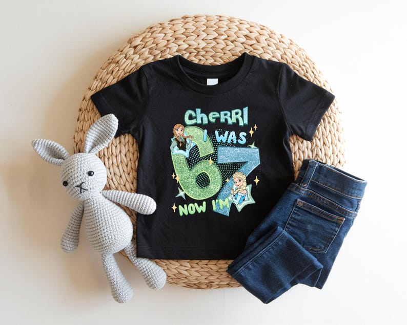 A black t-shirt with a graphic design, a gray crocheted rabbit, and a pair of blue jeans are arranged on a woven mat.
