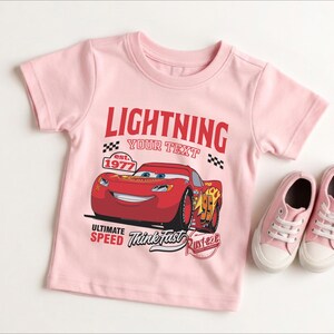 A pink t-shirt with a graphic of a red car and the text "LIGHTNING YOUR TEXT" printed on it, along with the text "EST. 1977" and "ULTIMATE SPEED Think fast Rusteze". The t-shirt is placed next to a pair of pink sneakers.