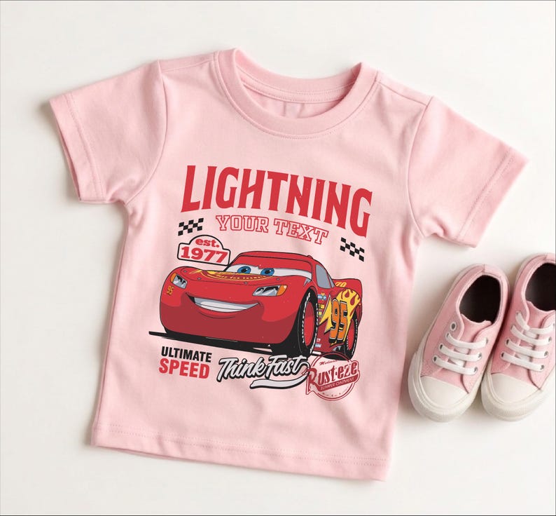 A pink t-shirt with a graphic of a red car and the text "LIGHTNING YOUR TEXT" printed on it, along with the text "EST. 1977" and "ULTIMATE SPEED Think fast Rusteze". The t-shirt is placed next to a pair of pink sneakers.