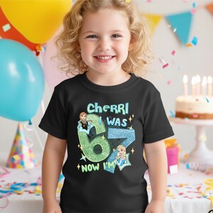 A young girl wearing a black t-shirt with a birthday message and a graphic of a character is smiling in front of a birthday cake with lit candles.