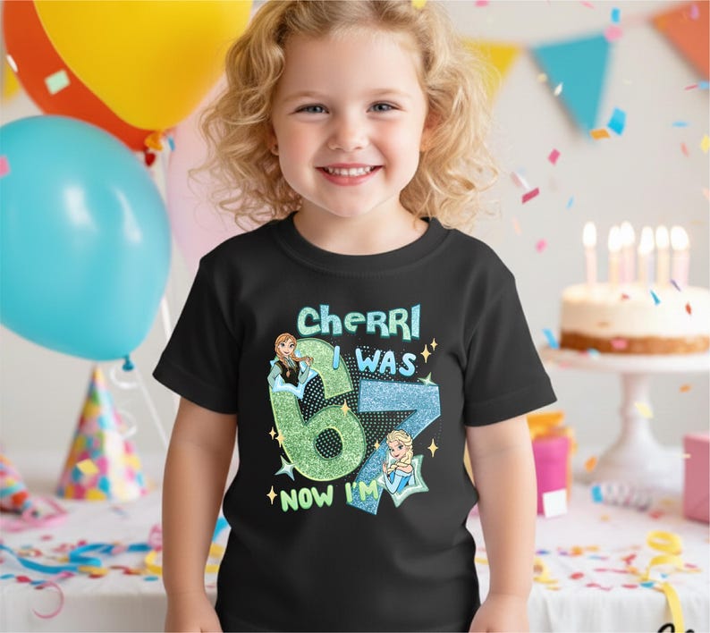 A young girl wearing a black t-shirt with a birthday message and a graphic of a character is smiling in front of a birthday cake with lit candles.