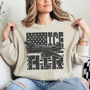 May include: A beige sweatshirt with a distressed graphic design featuring a vintage airplane flying over an American flag with the words "ICE FLYER" printed in a distressed font.