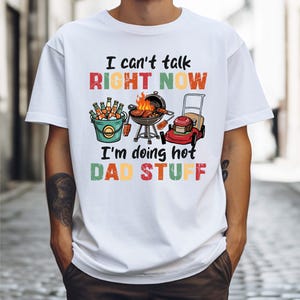 Può includere: T-shirt bianca con la scritta "I can't talk right now, I'm doing hot dad stuff." La grafica include un barbecue, un secchio di birra e un tosaerba.