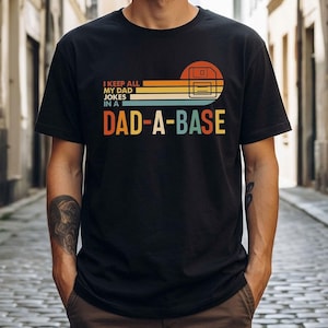 May include: Black t-shirt with a retro design featuring a floppy disk graphic and the text "I KEEP ALL MY DAD JOKES IN A DAD-A-BASE" in orange, yellow, and blue. The shirt has a crew neck and short sleeves.