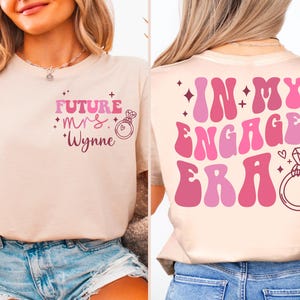 May include: A light pink t-shirt with the text "Future Mrs. Wynne" on the front and "In My Engaged Era" on the back. The text is in a pink and white retro font with a diamond ring graphic.