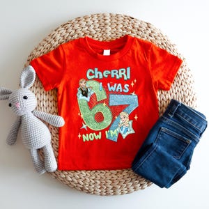 A red t-shirt with a cartoon character and text, a gray crocheted rabbit, and a pair of blue jeans are arranged on a woven basket.