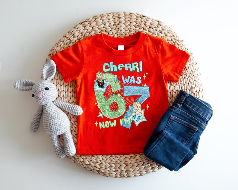 A red t-shirt with a cartoon character and text, a gray crocheted rabbit, and a pair of blue jeans are arranged on a woven basket.