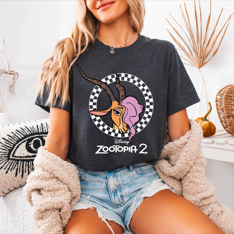 A woman wearing a black t-shirt with a graphic of a gazelle and the text "Zooopia 2" is sitting in a room with a decorative plant and a patterned pillow.