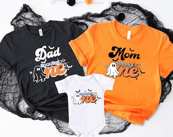 Puede incluir: Tres camisetas negras, naranjas y blancas con un gráfico de fantasma y el texto "Spooky One" en una fuente retro. Las camisetas son para una familia con un padre, una madre y un bebé.