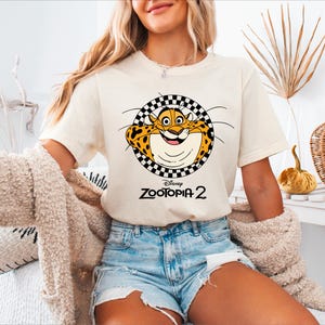 A woman wearing a white t-shirt with a cartoon leopard graphic and denim shorts, sitting in a room with a beige coat and a decorative plant.