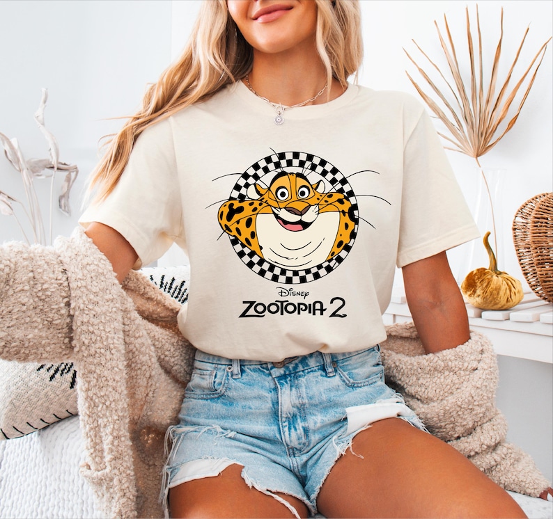 A woman wearing a white t-shirt with a cartoon leopard graphic and denim shorts, sitting in a room with a beige coat and a decorative plant.