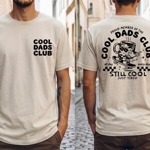 Könnte beinhalten: Beiges T-Shirt mit dem Text "COOL DADS CLUB" auf der Vorderseite und einer Grafik auf der Rückseite. Die Grafik auf der Rückseite zeigt eine Kaffeetasse auf einem Skateboard mit dem Text "PROUD MEMBER OF THE DADS CLUB" und "STILL COOL JUST TIRED".