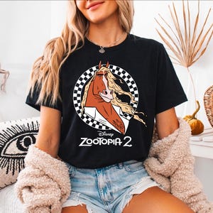 A woman wearing a black t-shirt with a graphic of a cartoon horse and the text "Disney Zootopia 2" is sitting in a room with a decorative plant and a patterned pillow.