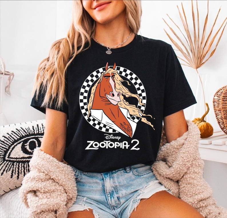 A woman wearing a black t-shirt with a graphic of a cartoon horse and the text "Disney Zootopia 2" is sitting in a room with a decorative plant and a patterned pillow.