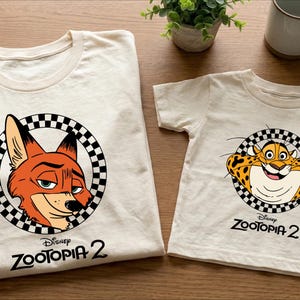 two white t-shirts with Disney Zootopia 2 logos, one for a larger size and one for a smaller size. The t-shirts are placed on a wooden surface, along with a coffee mug and a potted plant.