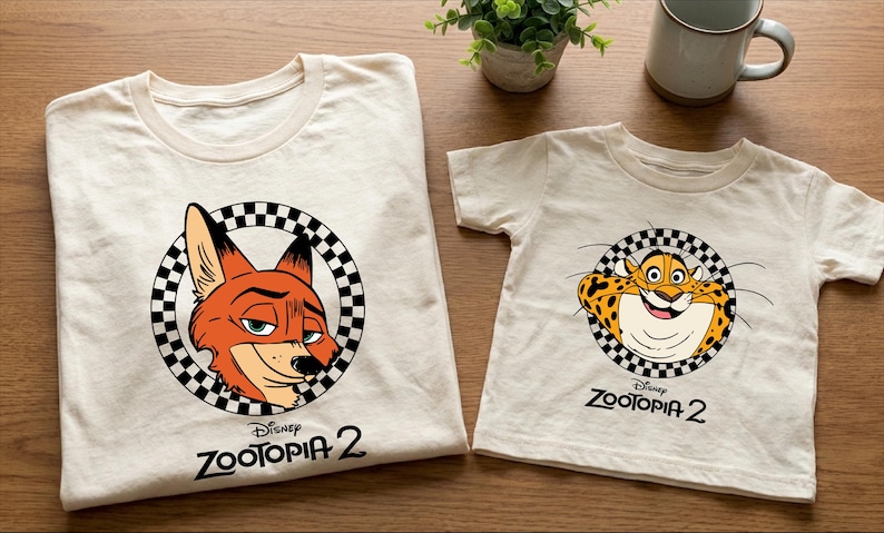 two white t-shirts with Disney Zootopia 2 logos, one for a larger size and one for a smaller size. The t-shirts are placed on a wooden surface, along with a coffee mug and a potted plant.