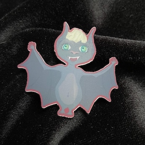 Könnte beinhalten: Ein Cartoon-Fledermaus-Aufkleber mit grauem Körper, roter Kontur und hellgrünen Augen. Die Fledermaus hat eine hellblonde Frisur und lächelt. Die Flügel sind ausgebreitet, auf schwarzem Hintergrund.