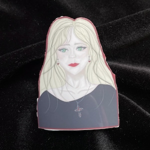 Könnte beinhalten: Ein Aufkleber mit einer stilisierten Illustration einer Frau mit langen blonden Haaren, grünen Augen und roten Lippen. Sie trägt ein schwarzes Oberteil und eine Kreuzkette. Der Aufkleber hat einen roten Rand.