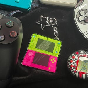 Puede incluir: Un llavero de consola de juegos portátil rosa brillante y verde con un dije plateado en forma de estrella. El llavero está rodeado de otros dispositivos de juego, incluido un controlador negro y una mascota digital retro.