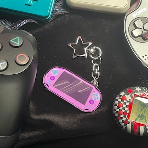Könnte beinhalten: Ein rosa Schlüsselanhänger in Form einer Handheld-Spielkonsole mit einem sternförmigen Anhänger. Der Schlüsselanhänger ist silberfarben und an einer kleinen Konsole mit lila Bildschirm und rosa Knöpfen befestigt. Andere Spielkonsolen sind im Hintergrund.