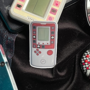 Puede incluir: Una pegatina de consola de juegos portátil roja y blanca con una pantalla en blanco, botones y estrellas y símbolos decorativos. Otras pegatinas similares son visibles en el fondo, sobre una superficie de terciopelo negro.
