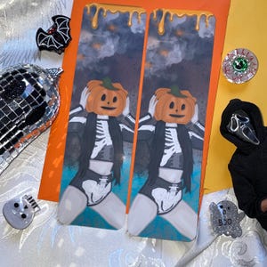 Könnte beinhalten: Zwei rechteckige Lesezeichen mit einer grafischen Illustration einer Figur mit Kürbiskopf und Skelett-Outfit vor einem dunklen, bewölkten Himmel mit orangefarbenen, tropfenden Akzenten an der oberen Kante jedes Lesezeichens.