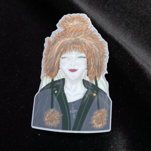 Könnte beinhalten: Ein Aufkleber mit einer stilisierten Illustration einer Person mit roten Haaren und einer schwarzen Jacke. Die Person zwinkert und trägt eine Halskette mit einem Kreuzanhänger. Die Jacke hat Fellakzente.