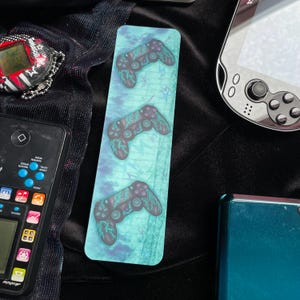 Könnte beinhalten: Ein Lesezeichen mit drei Gamecontroller-Designs mit türkisfarbenen Blitzen auf einem türkis-blauen Hintergrund. Das Lesezeichen ist rechteckig und hat eine glänzende Oberfläche. Weitere Retro-Gaming-Geräte befinden sich im Hintergrund.