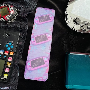 Könnte beinhalten: Lesezeichen mit einem sich wiederholenden Muster aus rosa Handheld-Spielkonsolen auf einem lavendelfarbenen Hintergrund mit Schneeflocken- und floralen Akzenten. Weitere Retro-Gaming-Geräte sind ebenfalls sichtbar.