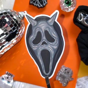 May include: A die-cut sticker featuring a gray, elongated ghost mask with two pointed horns at the top, set against a dark gray hood. The mask has large, hollow, black eye sockets and an open, elongated black mouth, creating a classic horror aesthetic.