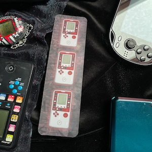 Könnte beinhalten: Eine Sammlung von Retro-Handheld-Gaming-Geräten und Zubehör. Das Bild zeigt einen Lesezeichen mit drei weißen und roten Spielkonsolen, ein schwarzes und rotes Tamagotchi, ein schwarzes Mobiltelefon und eine silberne Handheld-Konsole.