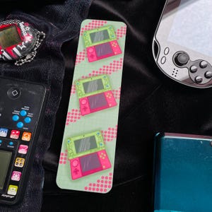 Könnte beinhalten: Ein Lesezeichen mit drei Illustrationen eines rosa und grünen Handheld-Spielgeräts. Das Lesezeichen hat einen hellgrünen Hintergrund mit rosa Punkten. Weitere Retro-Spielgeräte sind im Hintergrund zu sehen.