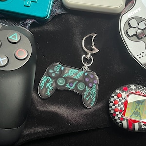 Könnte beinhalten: Schwarzer Anhänger in Form eines Controllers mit türkisblauem Blitzdesign und lila Knöpfen. Der Anhänger ist von verschiedenen Spielgeräten umgeben, darunter ein türkisblauer Controller, ein schwarzer Controller und eine Handheld-Konsole.