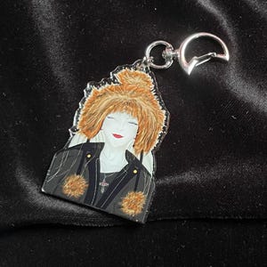 Könnte beinhalten: Ein klarer Acryl-Schlüsselanhänger mit einer stilisierten Illustration einer Frau mit roten Haaren und einer schwarzen Jacke. Der Schlüsselanhänger hat einen silbernen Verschluss und einen silbernen Schlüsselring. Die Frau trägt eine Kreuzkette.
